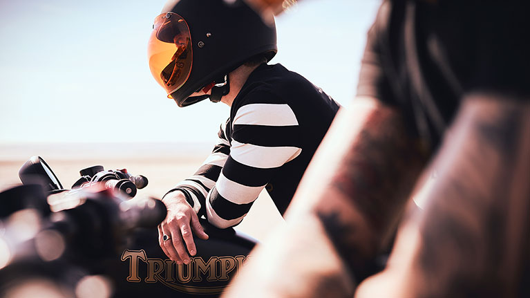 Male model wearing Triumph SS20 lifestyle clothing collection sitting on a Triumph Bonneville Bobber Black on a beach in summer
