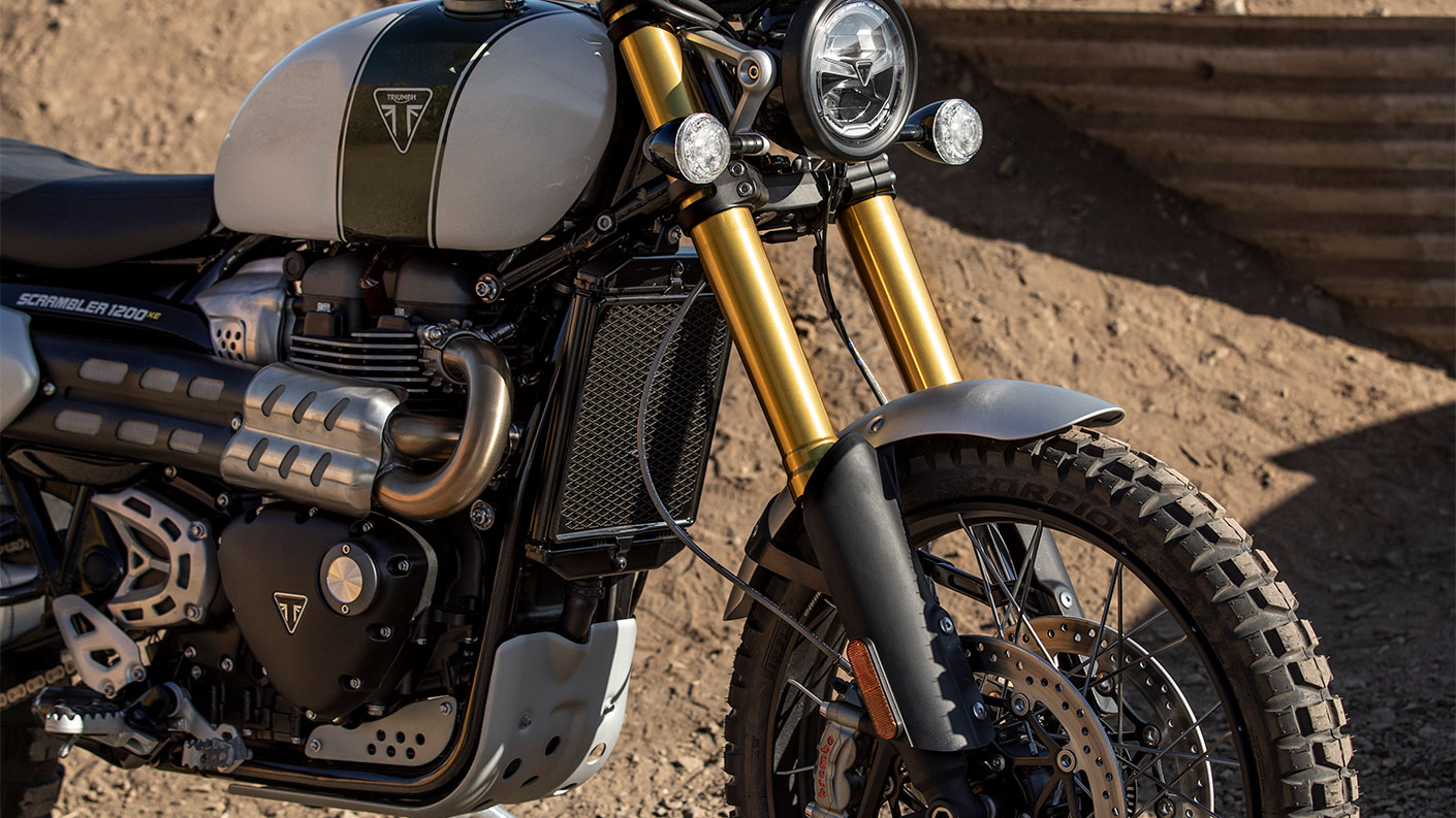 Triumph Scrambler 1200 XE stationary with sunlight illuminating gold front forks, sculpted exhaust and Triumph badging