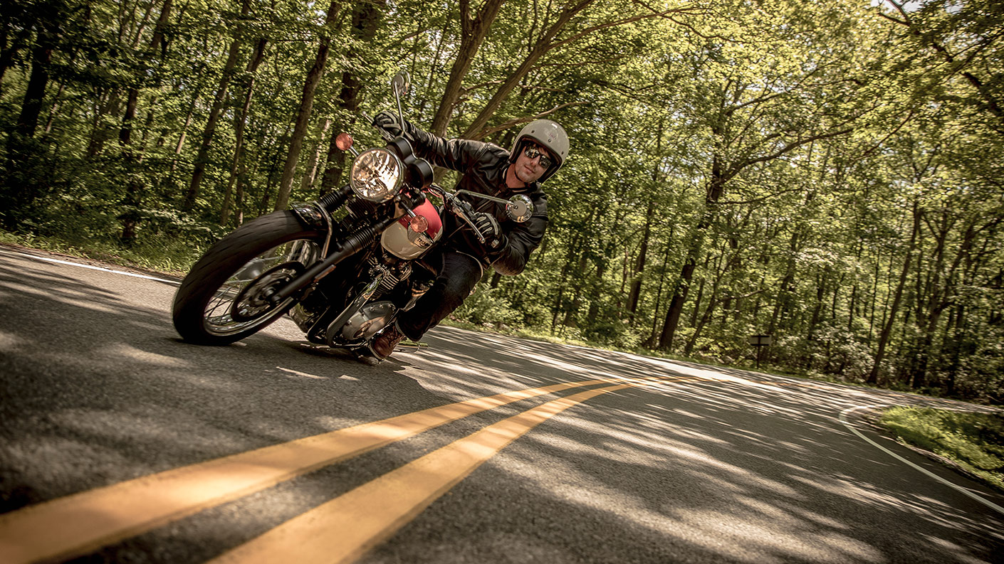 Triumph Bonneville T100 Motorcycle in white and orange variant cornering on a woodlands road.